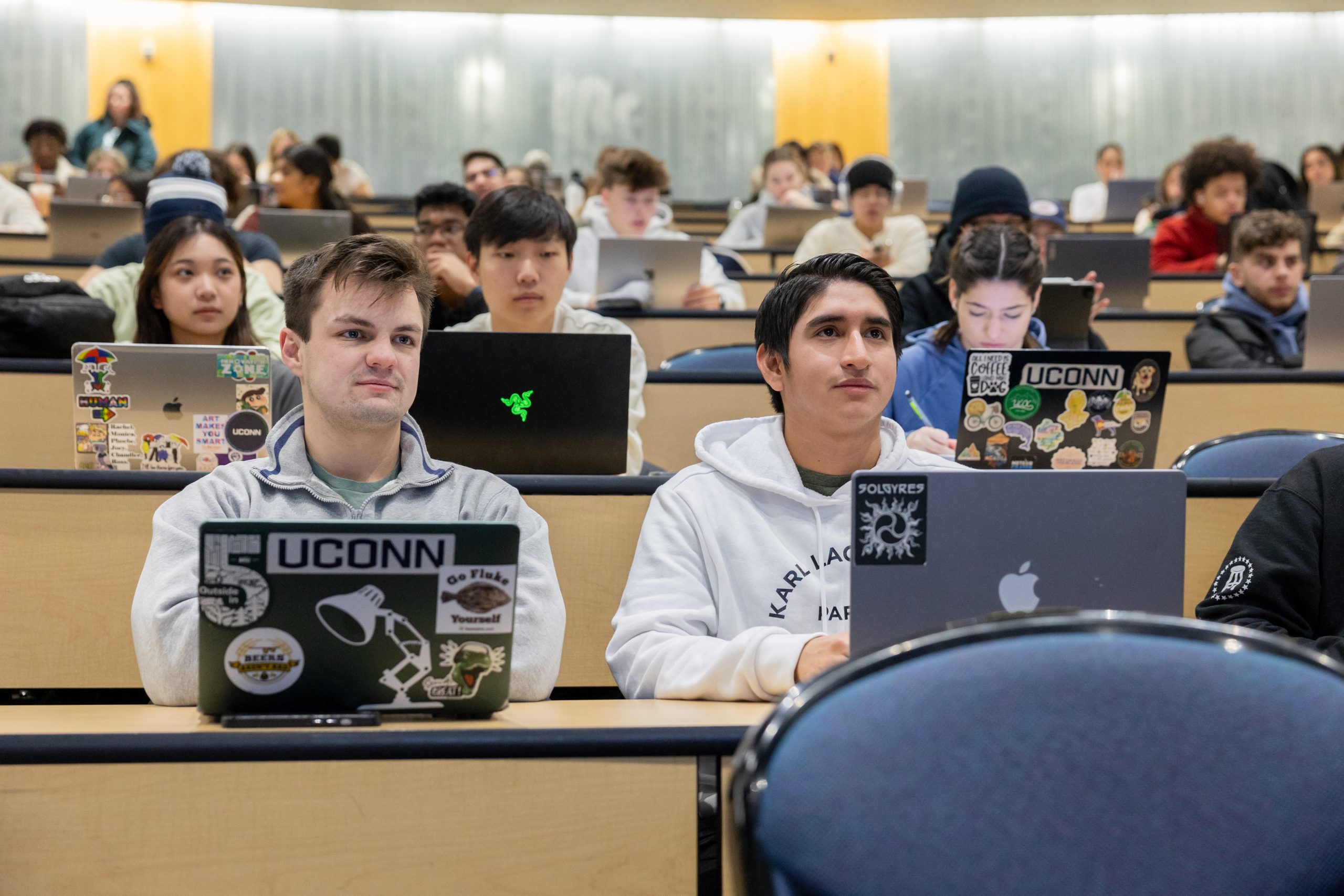 Rows of male and female students representing diverse backgrounds sit facing the camera in a large lecture hall, some looking up at their instructor, some writing or typing, all with laptops opened in front of them - Laptops in the first two rows are adorned with a variety of stickers, including three with prominent "UCONN" wordmarks.