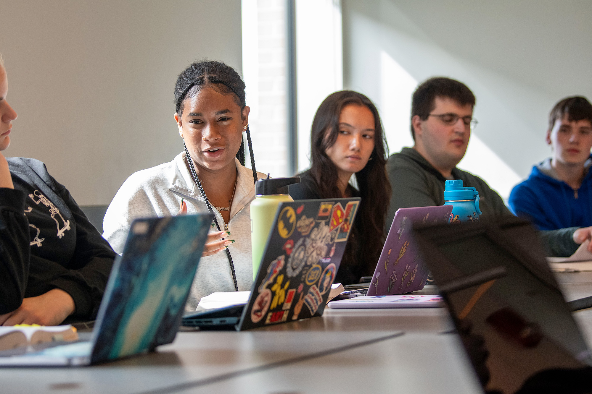 A Black female student wearing her hair in braids gestures with her hand while speaking to someone near the camera, while sitting at a table flanked by four other students, all with laptops opened in front of them