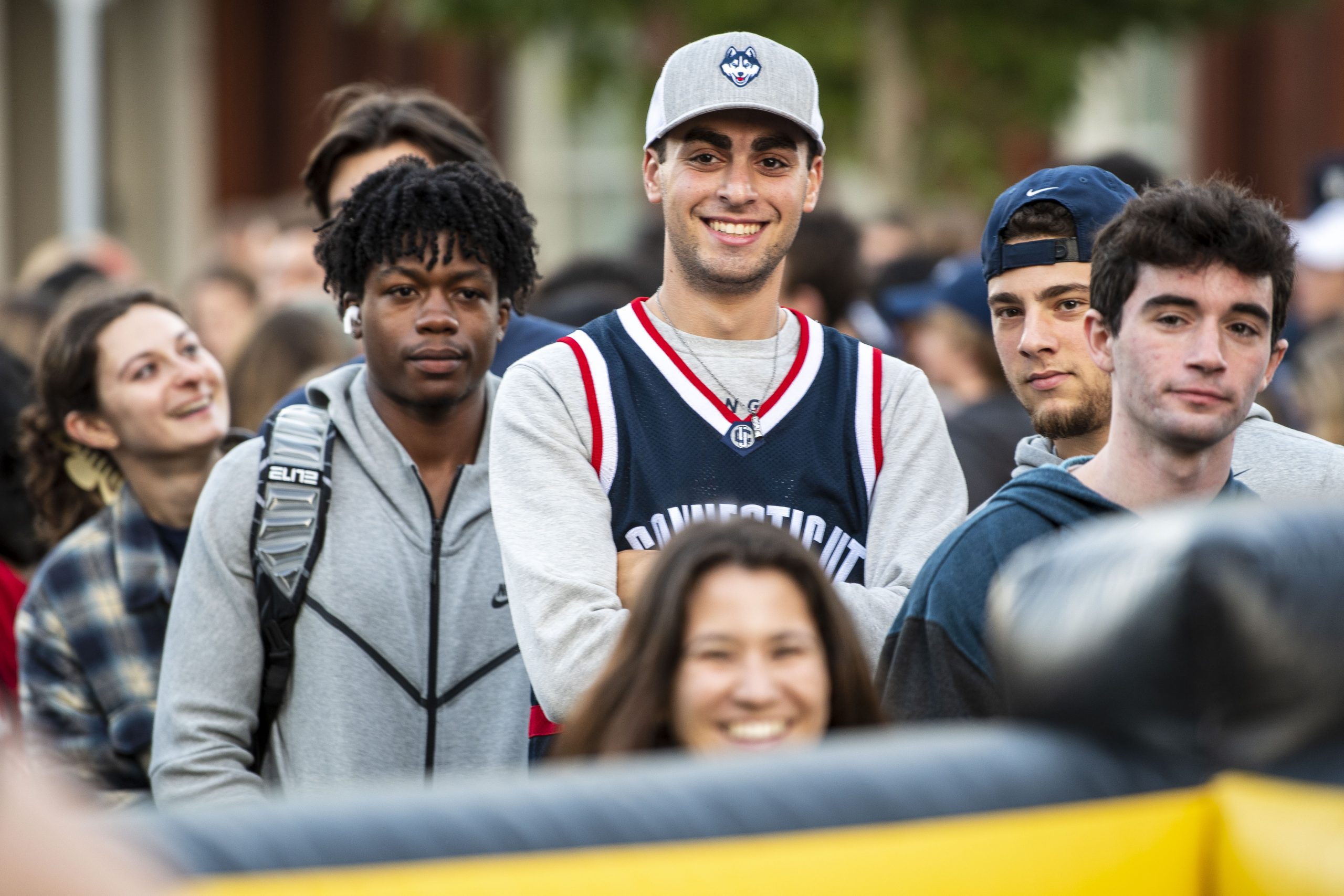 Two female and five male students are among a large group of students gathered outside Gampel Pavilion before First Night. The male student at the center smiles and is wearing a UConn Husky logo hat and a "Connecticut" basketball jersey.