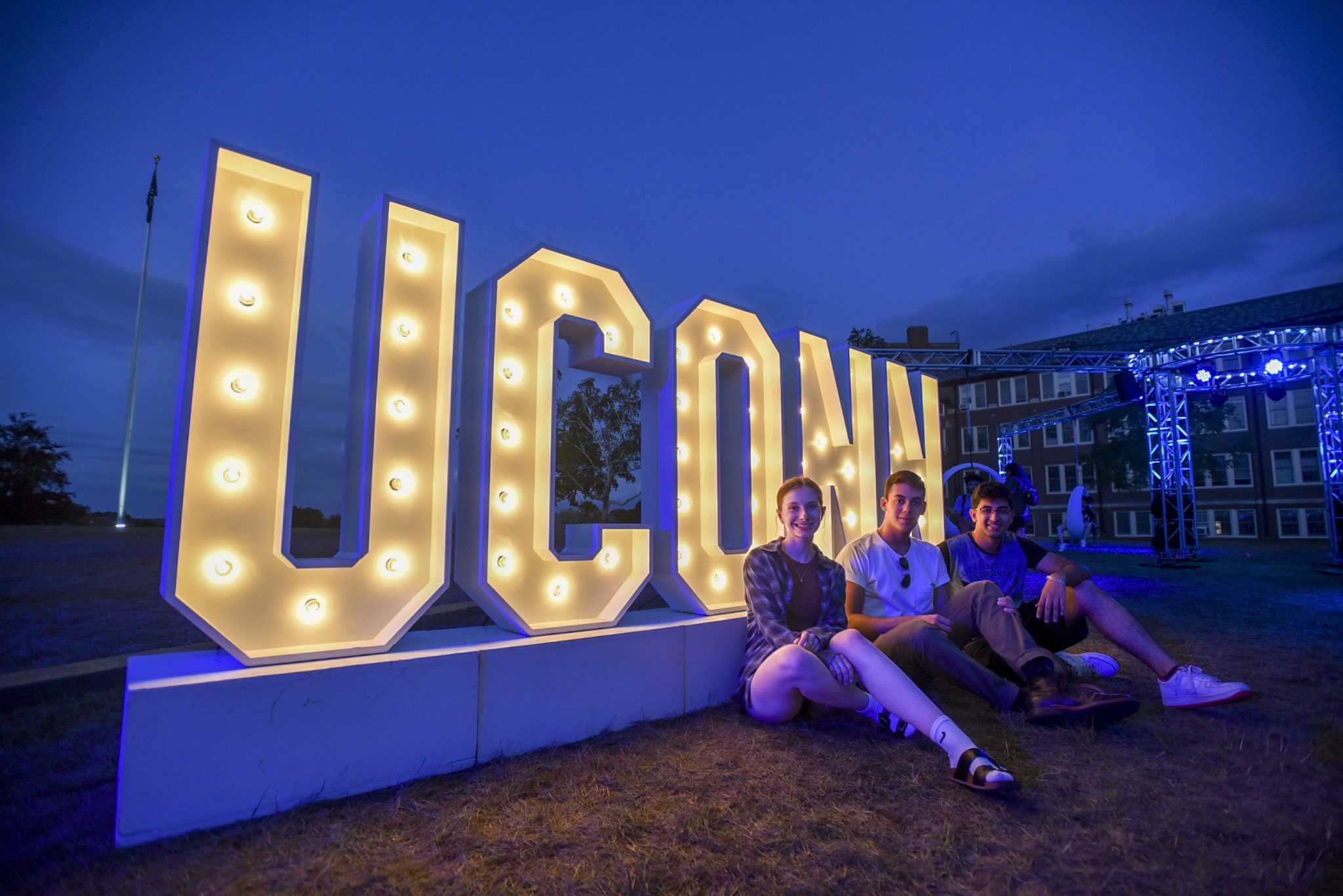 A female and two male students sit on the ground, smiling at the camera, in front of a lighted "UCONN" sign at dusk, with a dark blue sky above and blue lights shining from a large metal rig in the background.