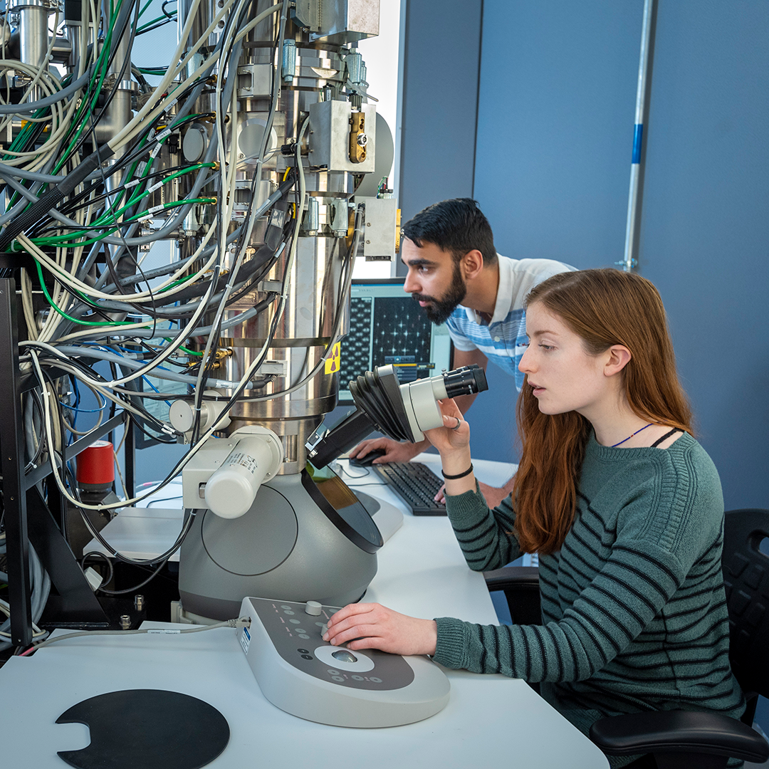 A white female student looks into the lens of an electron microscope with a male student of color behind her, looking at a computer screen. Dozens of wires feed into the large metal cylinder of the microscope, taking up more than half the image.