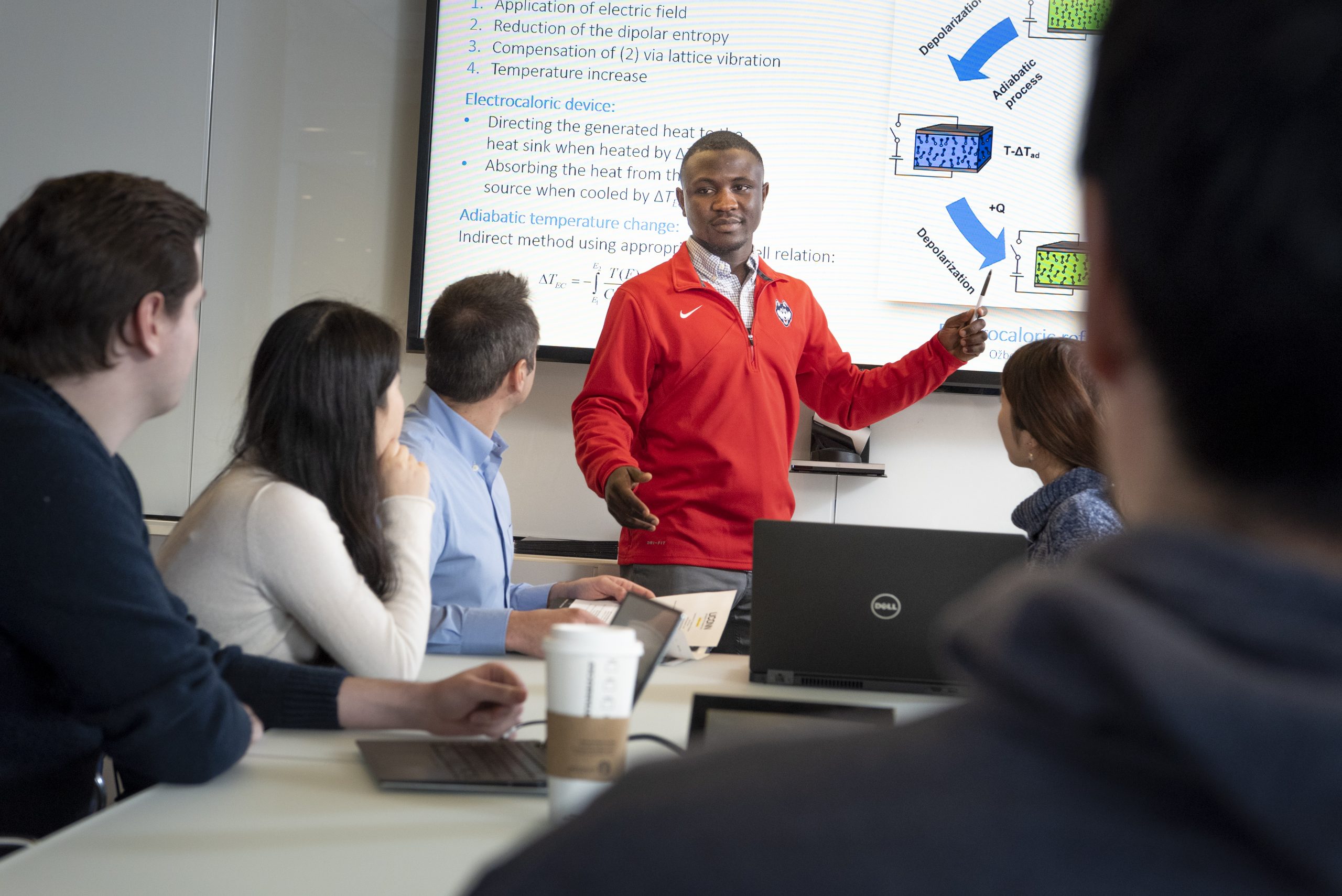 A Black male wearing a red pullover featuring the UConn Husky dog logo points with a pen to a scientific presentation on a screen behind him while presenting to a group of males and females who are all facing him.