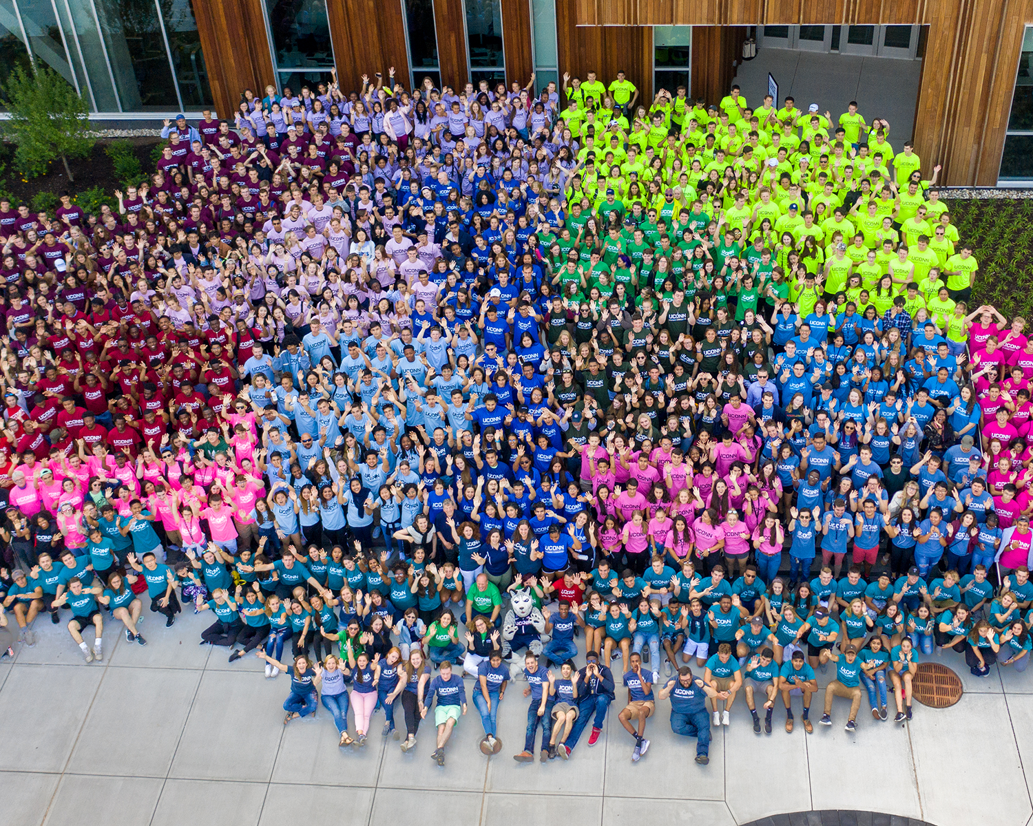 Hundreds of diverse students pose together, many waving at the camera and smiling and grouped into about a dozen sections by matching color-coded shirts that indicate which UConn Learning Community they belong to.