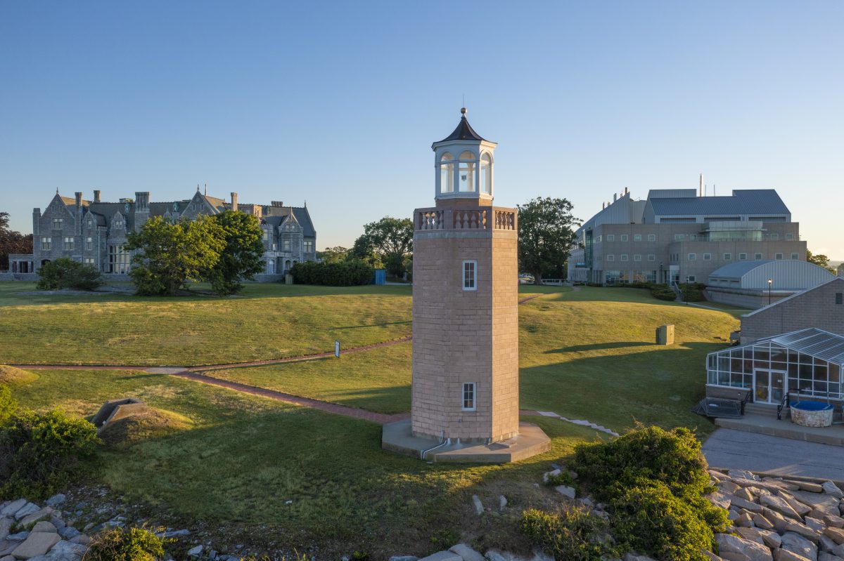The Avery Point Lighthouse, a concrete structure with a large wooden lantern at the top, flanked by the Branford House - a large stone structure - and the Marine Sciences building