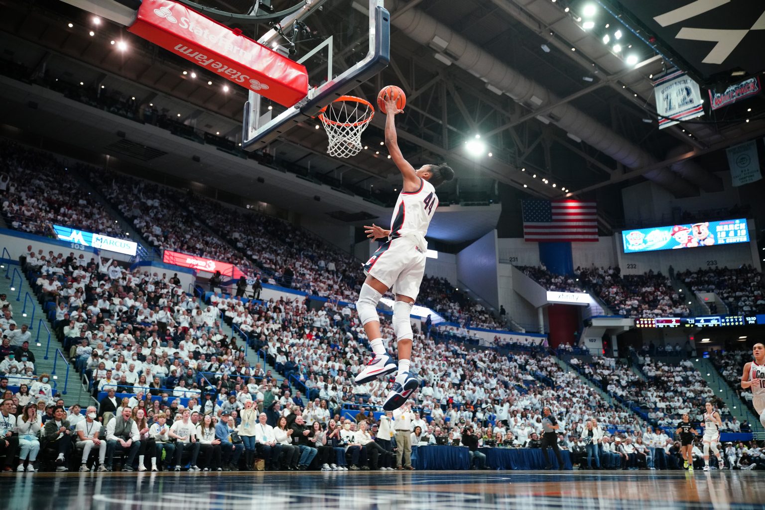 A Black woman's basketball player wearing a #44 UConn jersey jumps to make a shot into the opposing team's basketball hoop in a large arena with a crowd full of fans wearing white shirts, many with their mouths open expressing awe and excitement