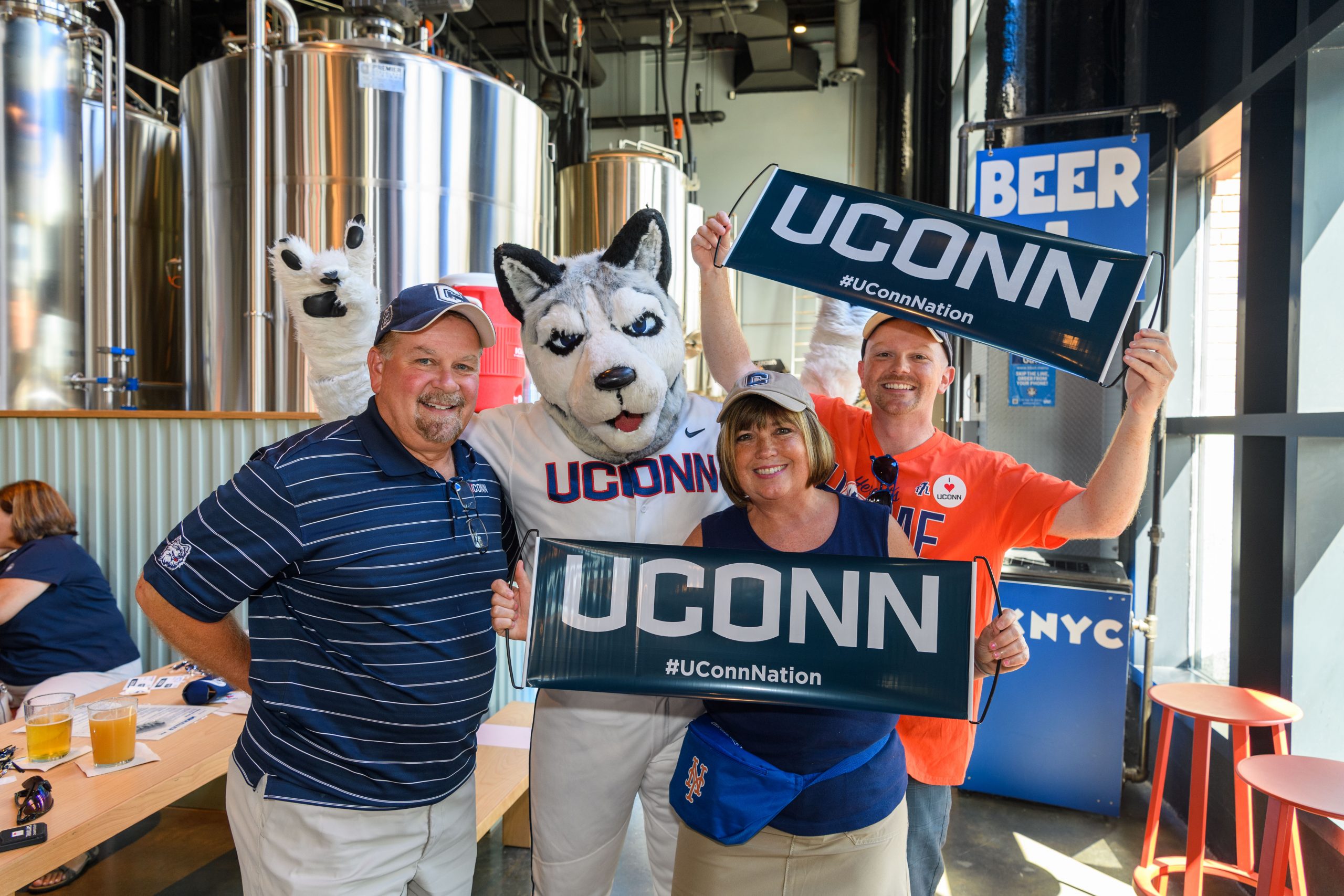 The costumed Jonathan the Husky mascot, wearing a baseball uniform, poses for a photo with three middle-aged UConn fans holding UConn banners at in front of large metal beer brewing vats at an alumni event before a New York Mets baseball game.