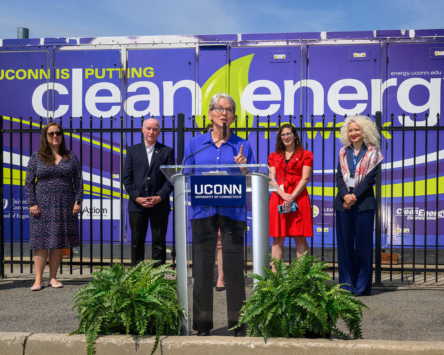 U.S. Secretary of Energy Jennifer Granholm speaks into a microphone at a clear podium with her left index finger pointing upward during a press conference at the Center for Clean Energy Engineering on May 20, 2022. Behind Granholm, four people look on, smiling, including U.S. Rep Joe Courtney and University President Radenka Maric. Behind them is a graphic on a fuel cell that says "UConn is putting clean energy to work."