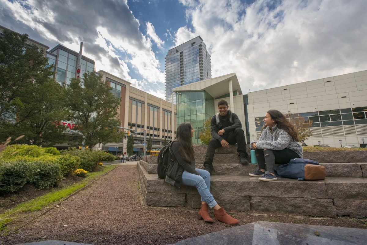 A male student and two female students representing various ethnic backgrounds sit facing each other, smiling and talking, on a stone wall outside UConn Stamford with city buildings in the background