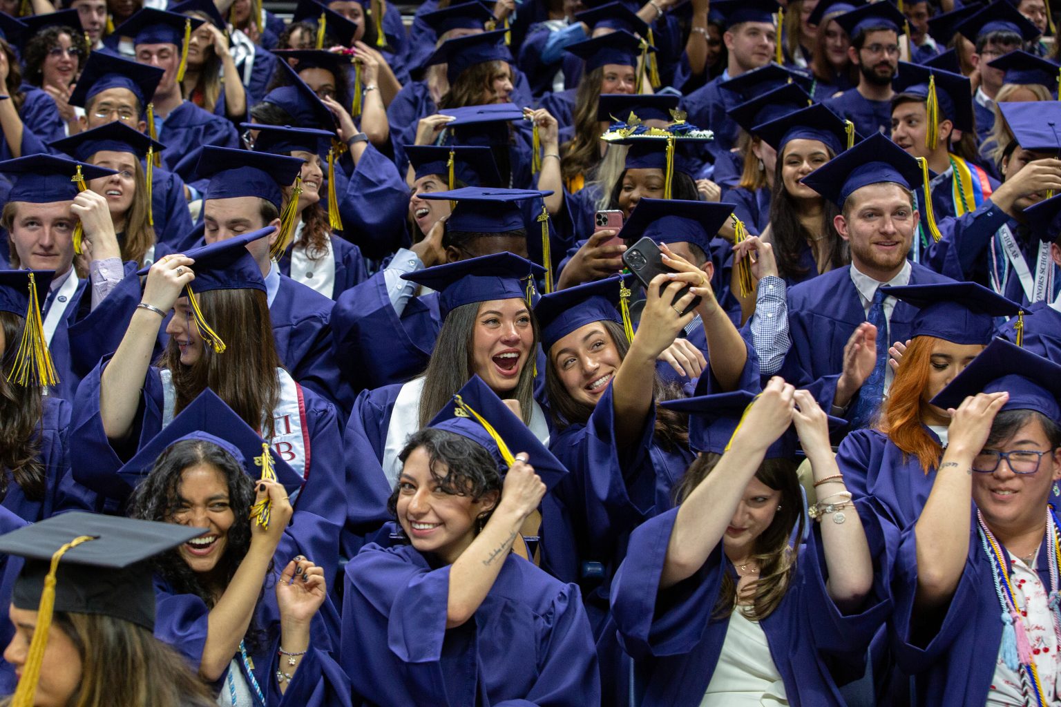 A group of students wearing blue graduation caps and gowns and representing various ethnic backgrounds and genders joyfully move the tassels on their caps at the College of Liberal Arts and Sciences (CLAS) Commencement ceremony. Many smile, some pose for selfies.