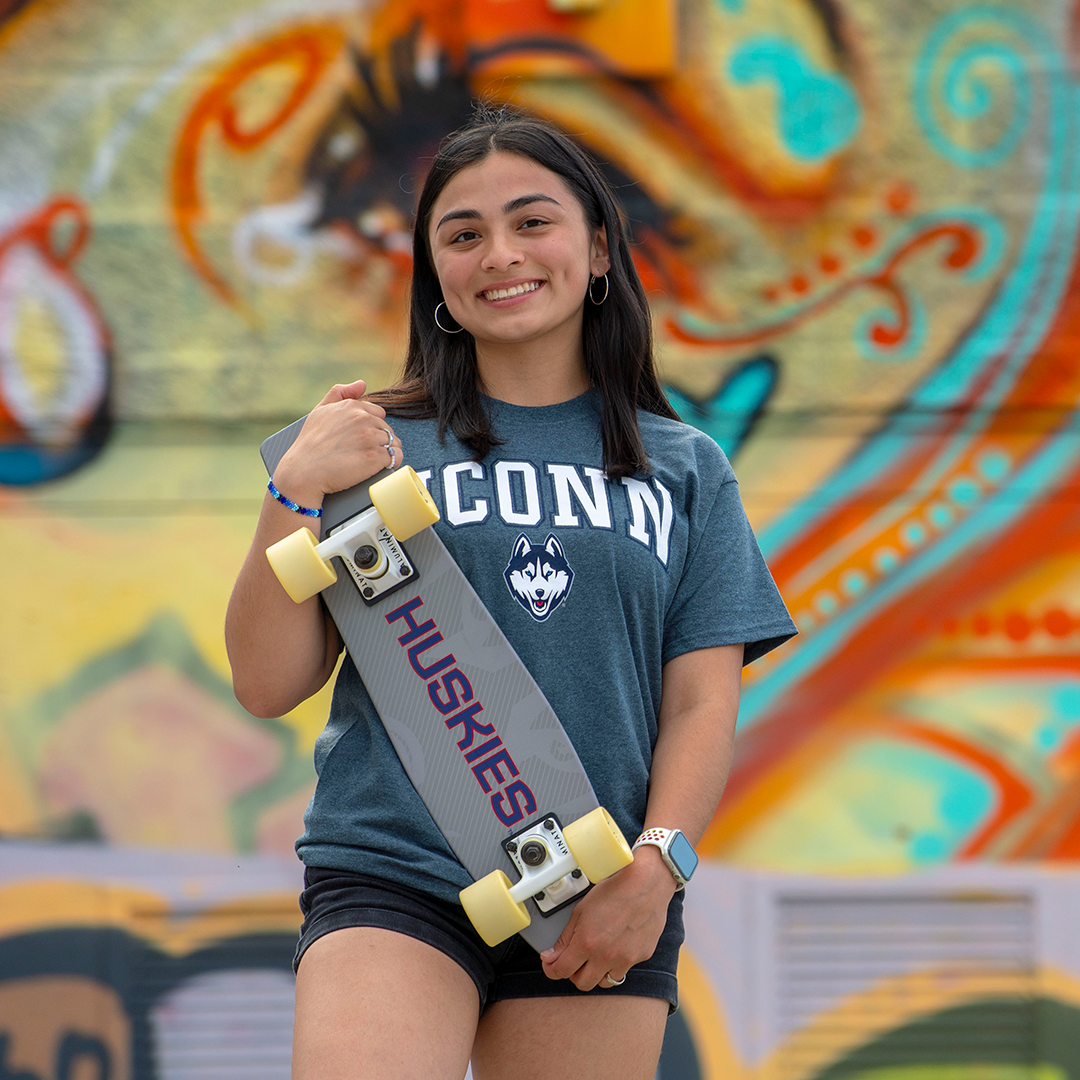 A female UConn Hartford student with dark hair smiles at the camera, wearing a "UConn" t-shirt with the Husky dog logo and holding a skateboard, wheels out, that says "Huskies" on the bottom, in front of a colorful mural at Heaven Skate Park in Hartford.