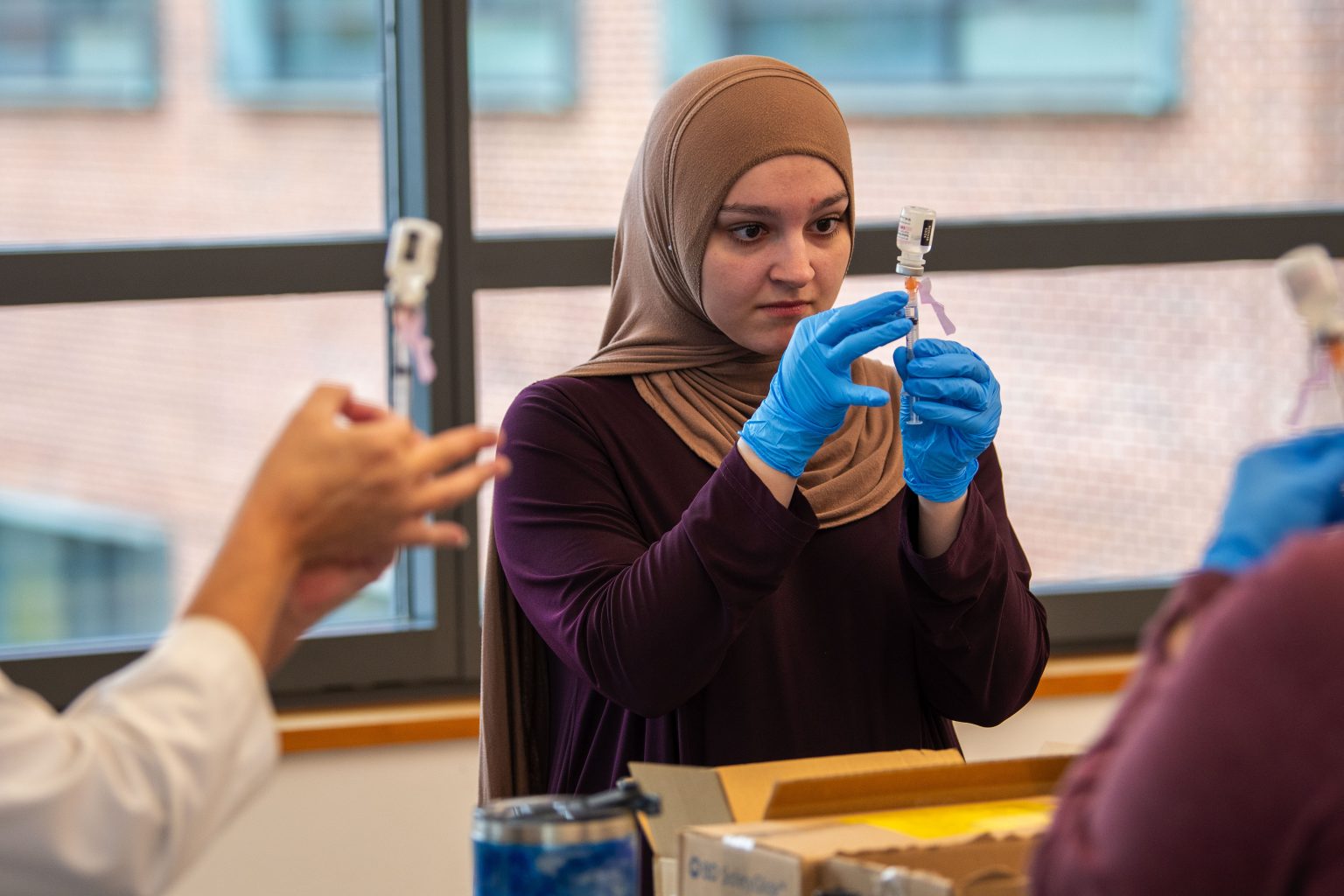 A female student wearing a hijab and blue latex gloves holds a needle vertically, drawing a liquid from a small glass vial, while the hands of two other people are seen doing the same on the edges of the frame.