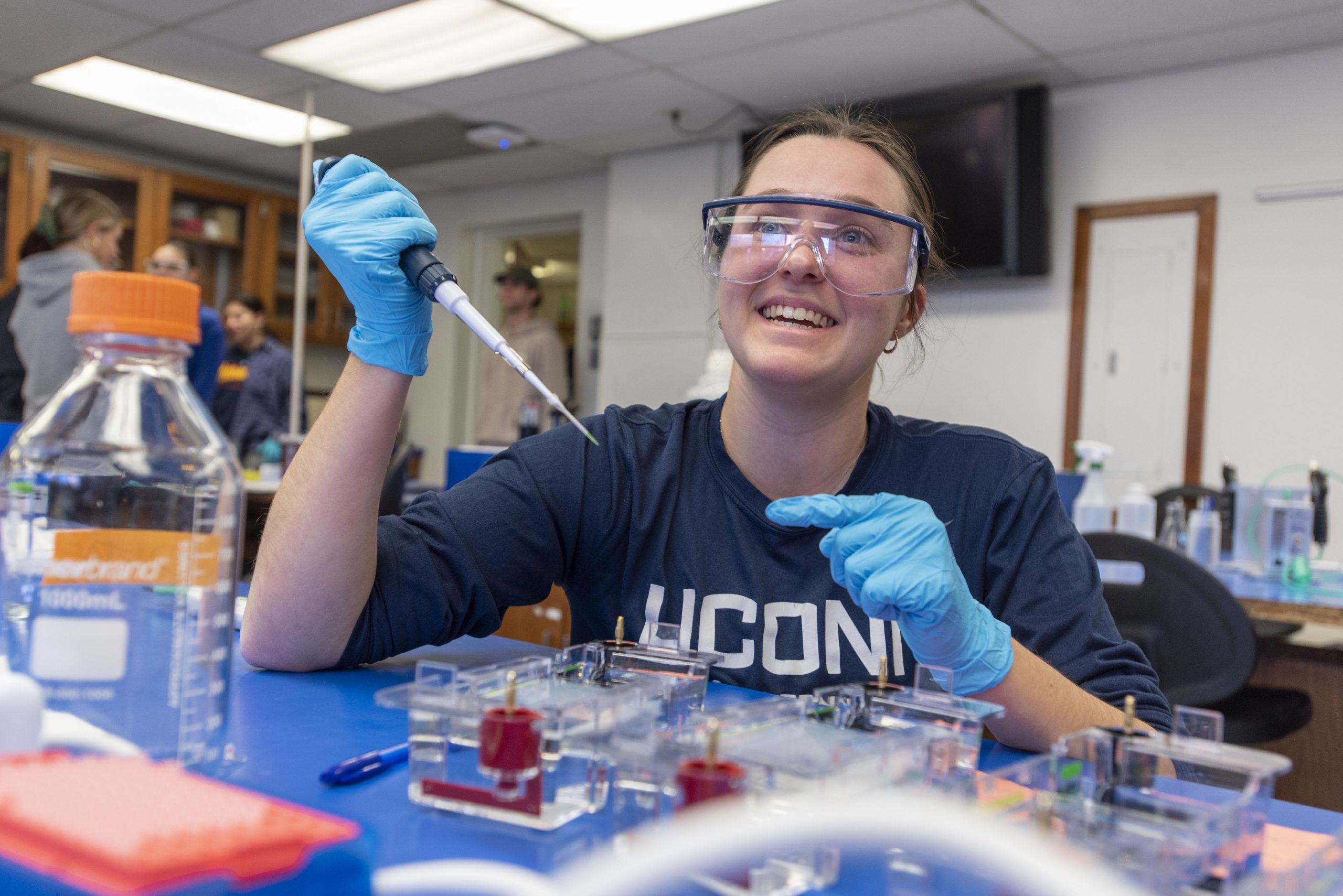 A white female college student wearing a blue shirt that says "UConn," blue latex gloves, and safety glasses holds a large pipette and smiles at someone just to the left of the camera in a lab classroom.