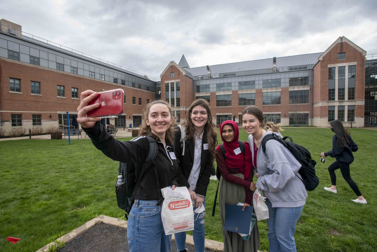 Four female students pose together, smiling, for a selfie in the courtyard in front of UConn Waterbury's main building.