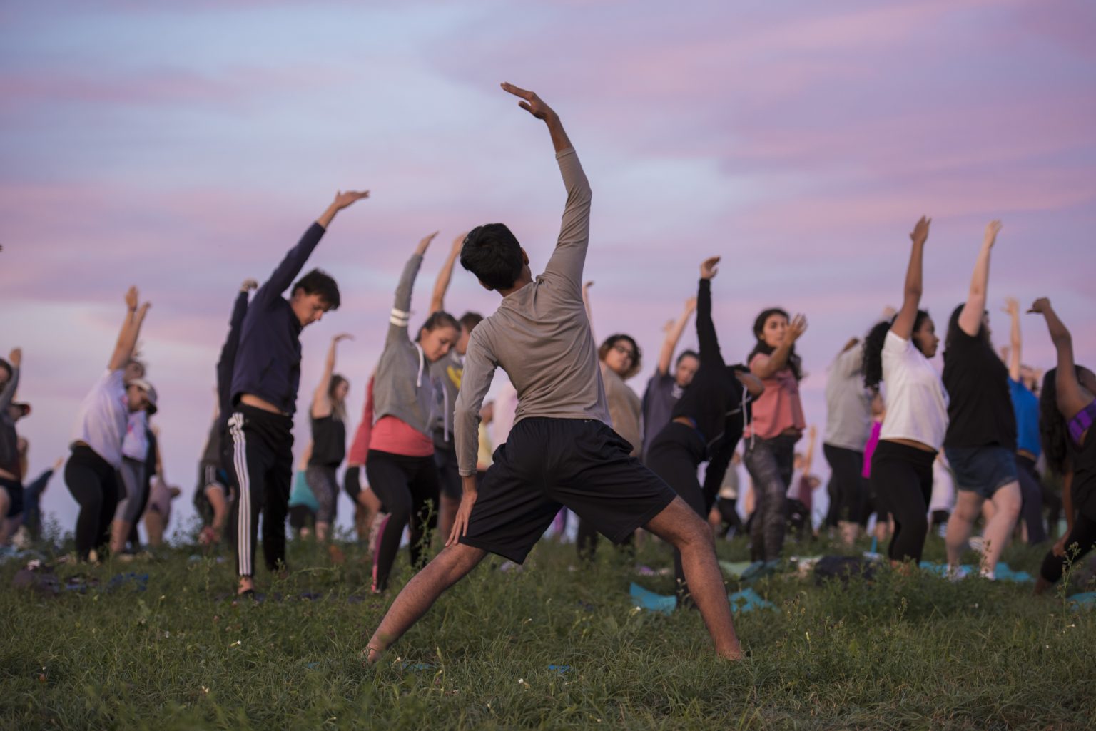 In the foreground, a male student of color stands in the grass and faces away from the camera in a yoga pose with his left arm on his left thigh and his right arm curved overhead. In the background, a diverse group of both male and female students attending the Yoga on Horsebarn Hill event face the camera, getting into the same pose.